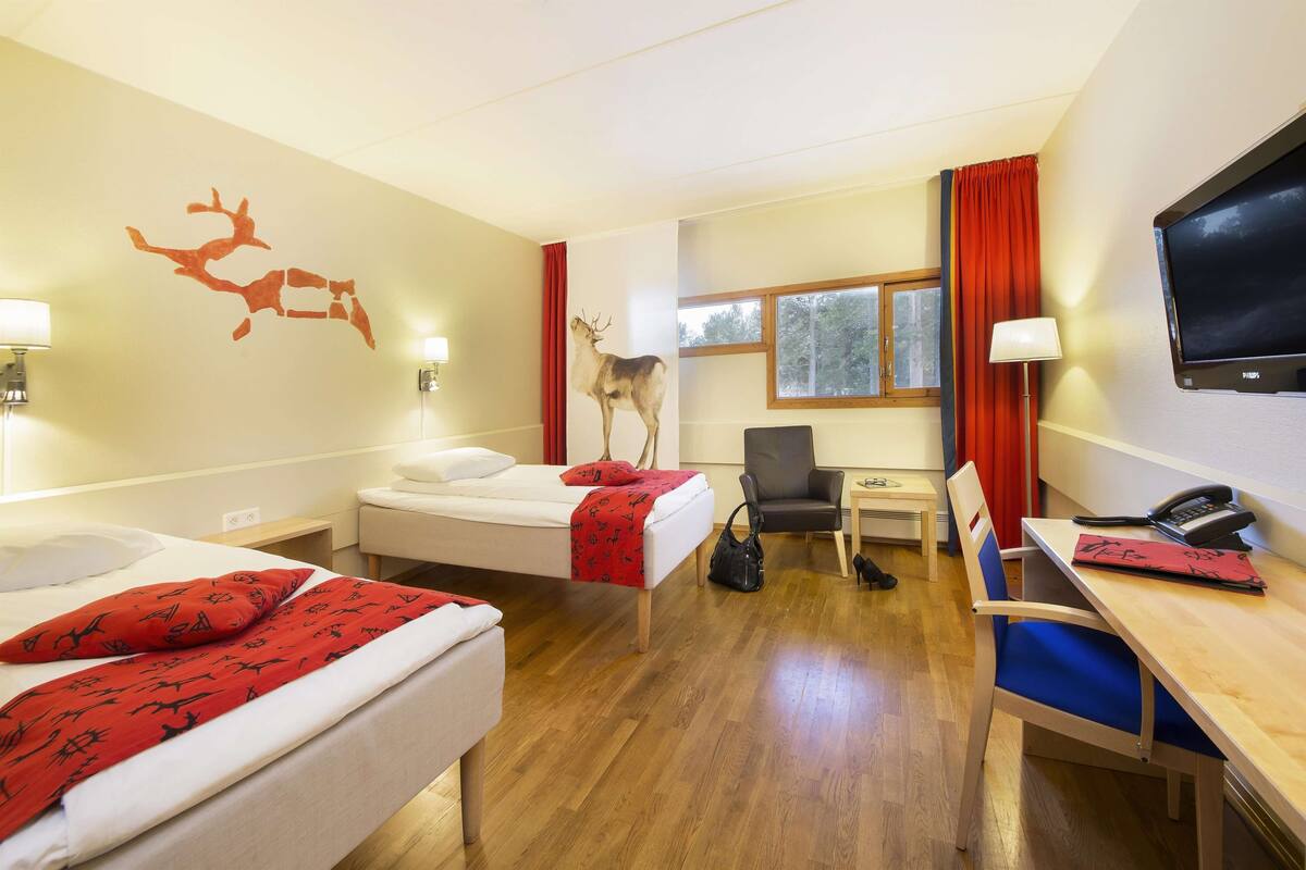 A hotel room with two beds, a desk, a chair, a TV, and a window.