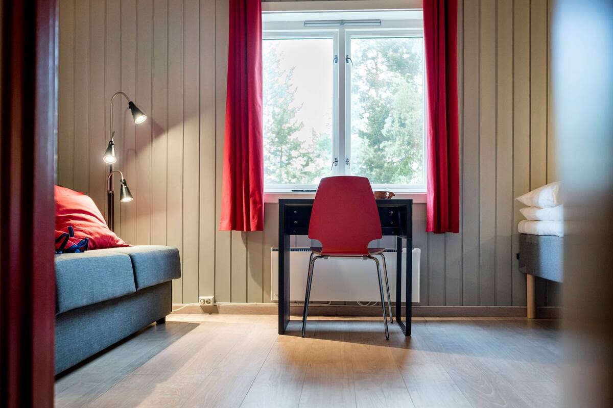 A room with a grey sofa, a red chair, a desk, and a window with red curtains.