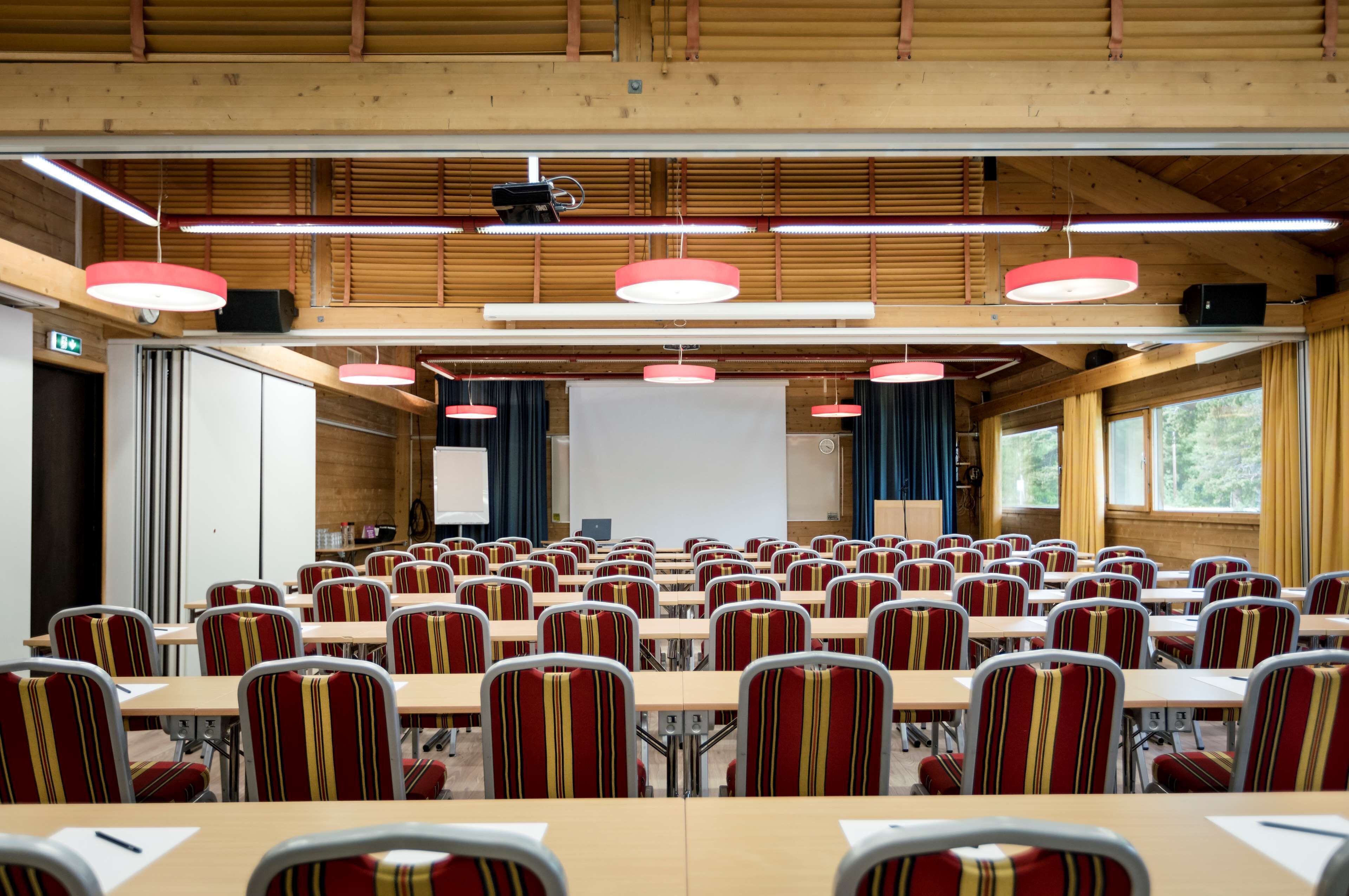A conference room with rows of tables and chairs, a projector screen, and a large window with curtains.