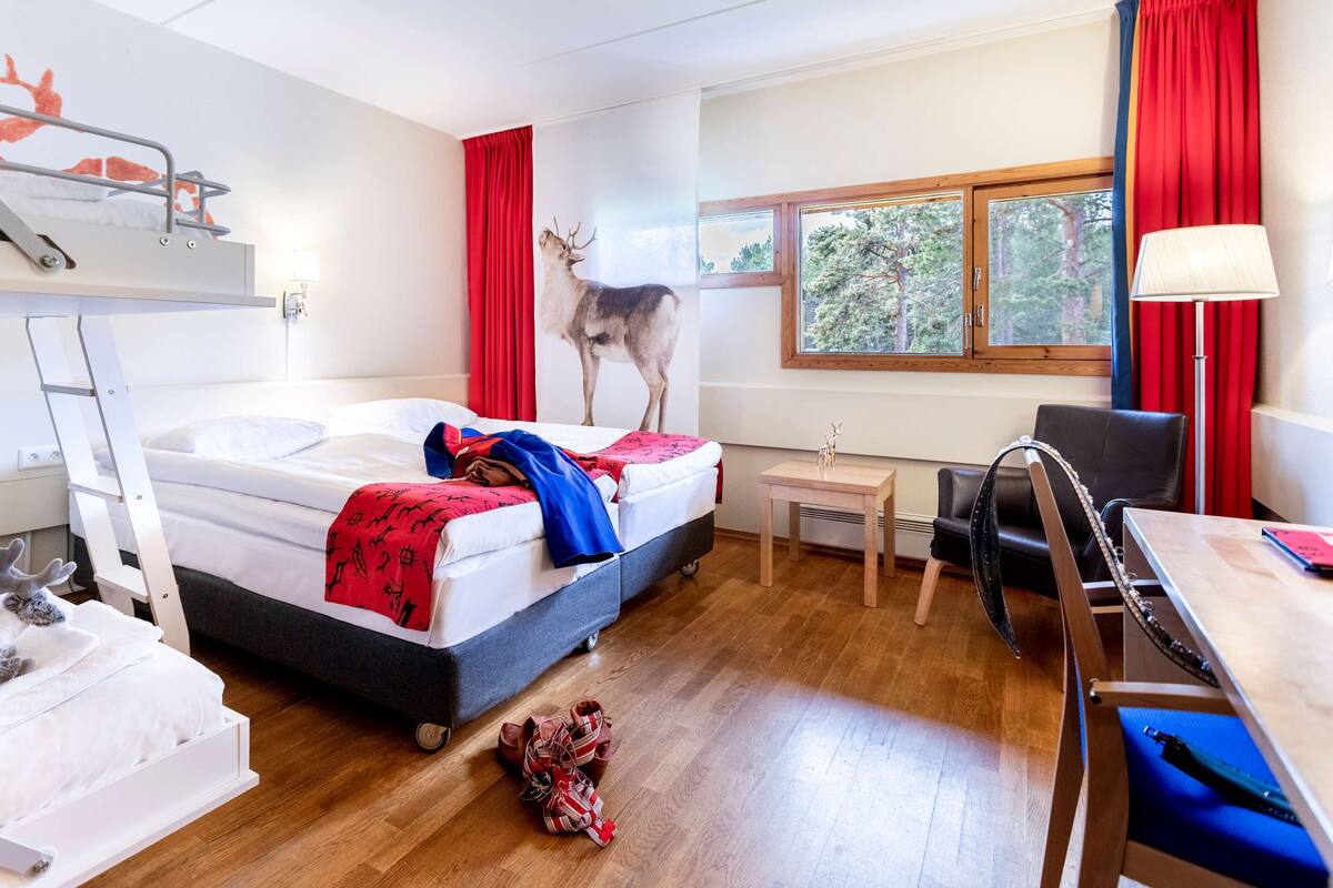 A hotel room with a bunk bed, a desk, a chair, a small table, and a window with a view of trees.
