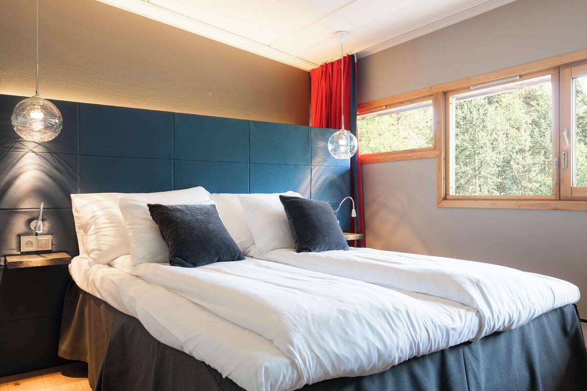 A modern bedroom with a large bed, a nightstand, a wall-mounted lamp, and a window with a view of trees.