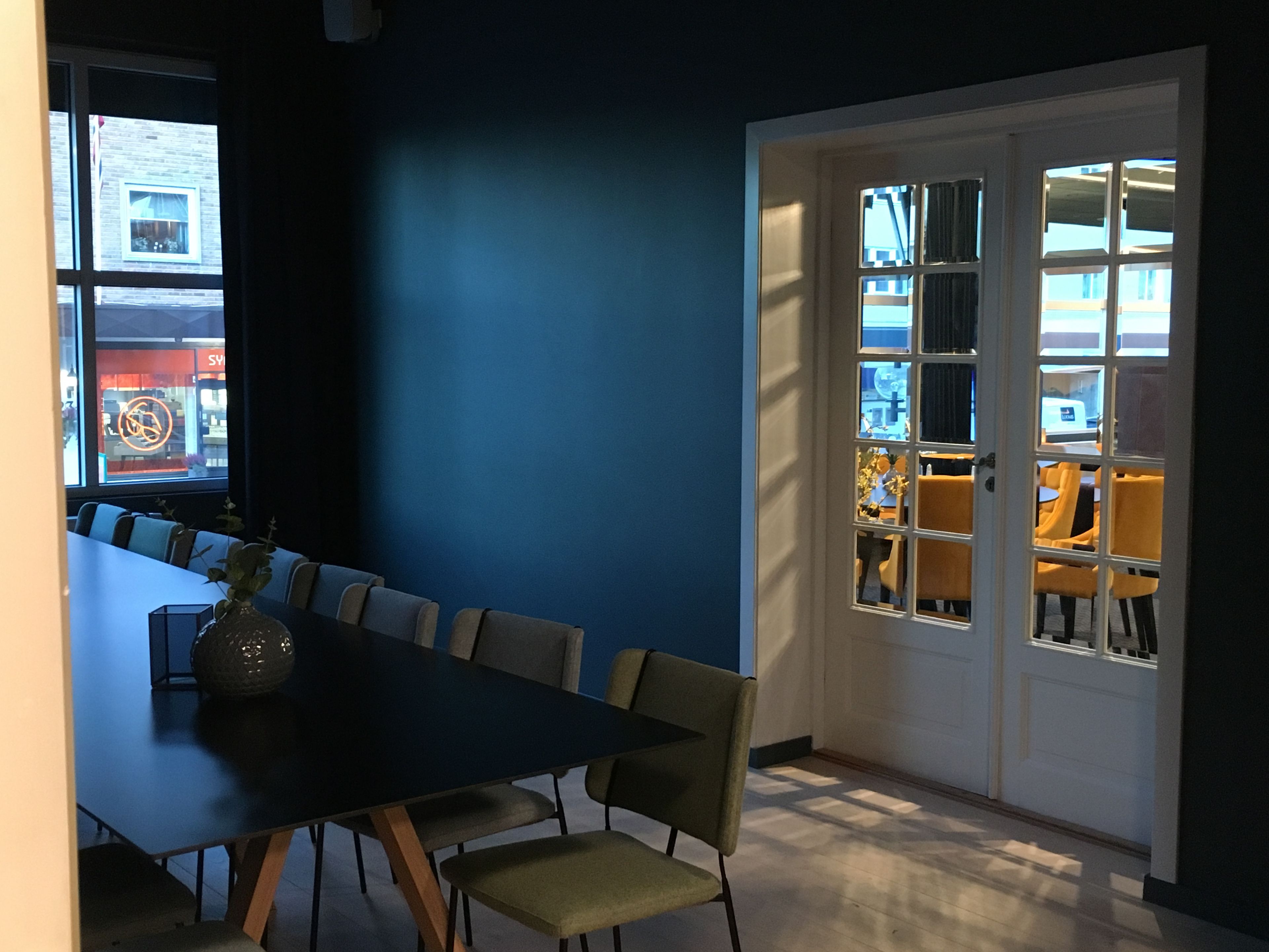 A conference room with a long table and chairs, a vase on the table, and a view through a door to an outdoor seating area.