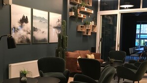 A modern living room with a brown sofa, green armchairs, and a small table with a plant. There are framed pictures on the wall and a cactus plant.