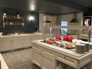 Montaje de bufé con diferentes alimentos y utensilios, muebles blancos y pared oscura con estantes.
