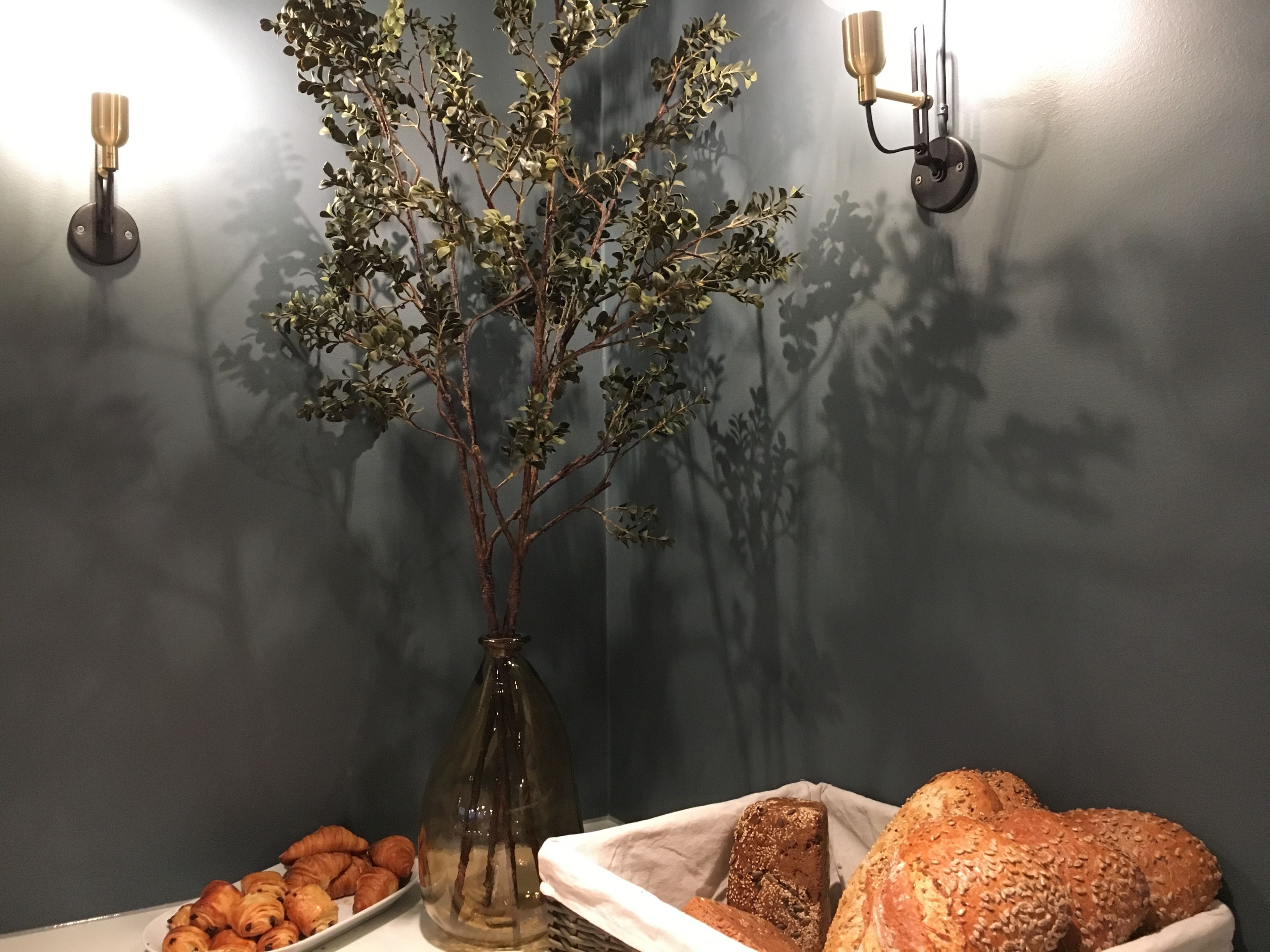 A table with pastries and bread, a vase with branches, and wall-mounted lights against a dark wall.