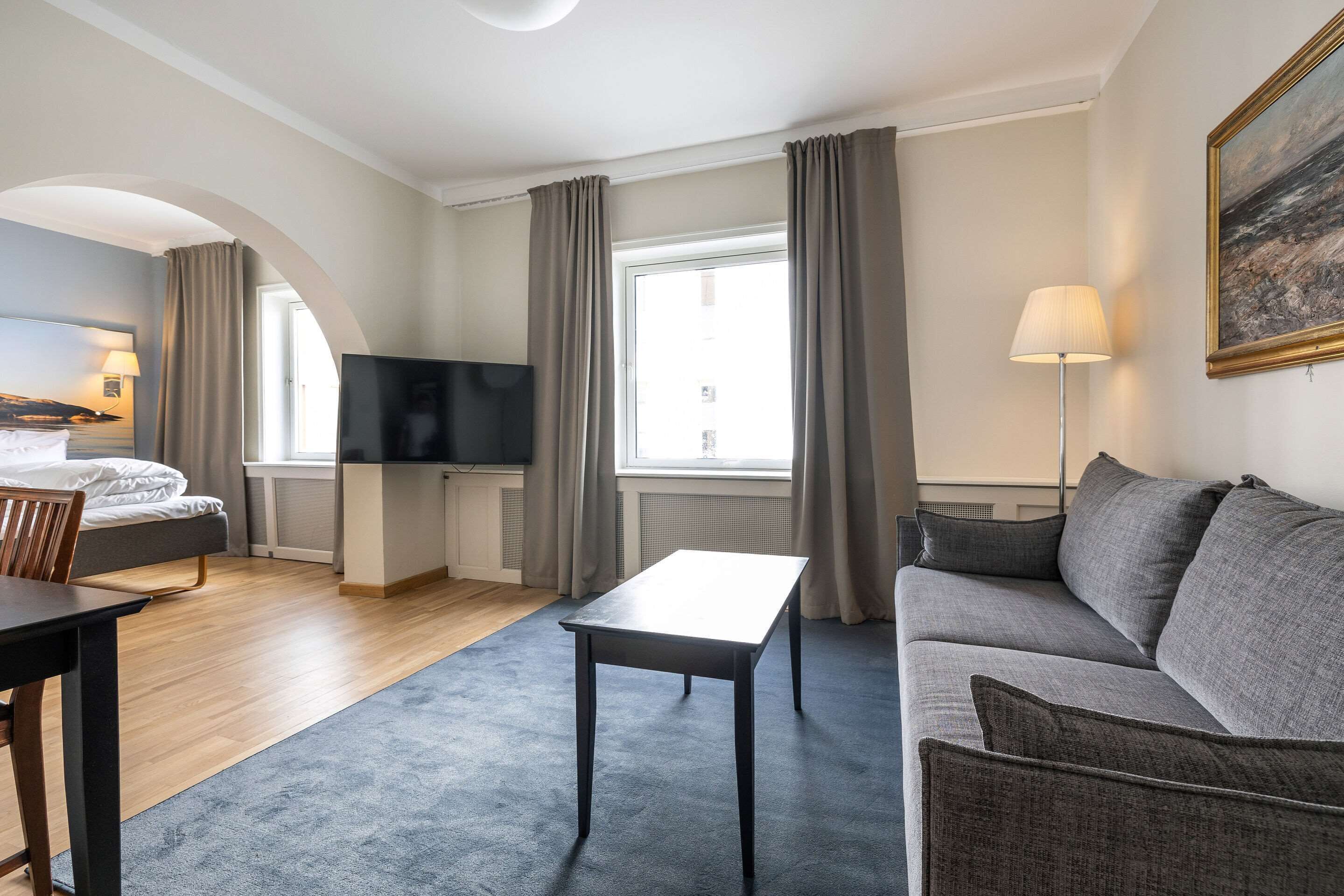 Quarto de hotel com cama, sofá, mesa, televisão e janela com cortinas.