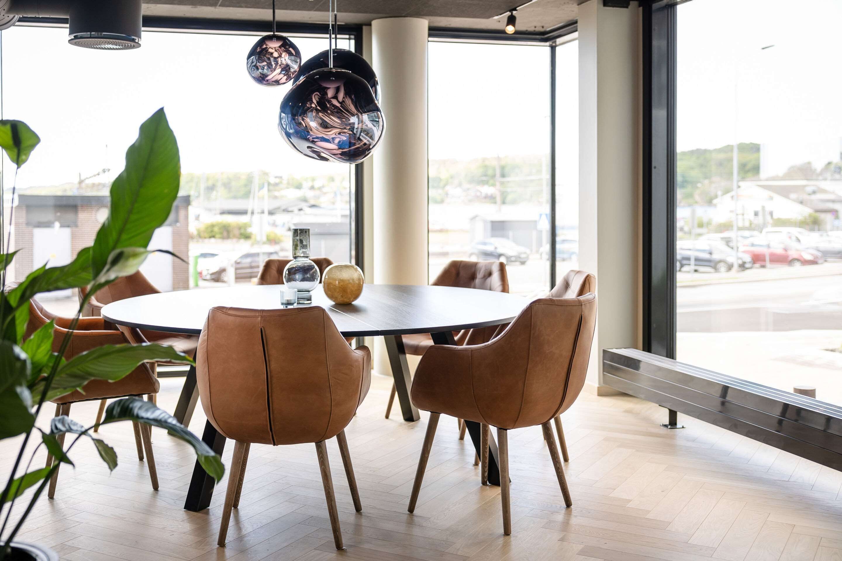 A modern dining area with a round table, brown chairs, and large windows offering a view of the street.