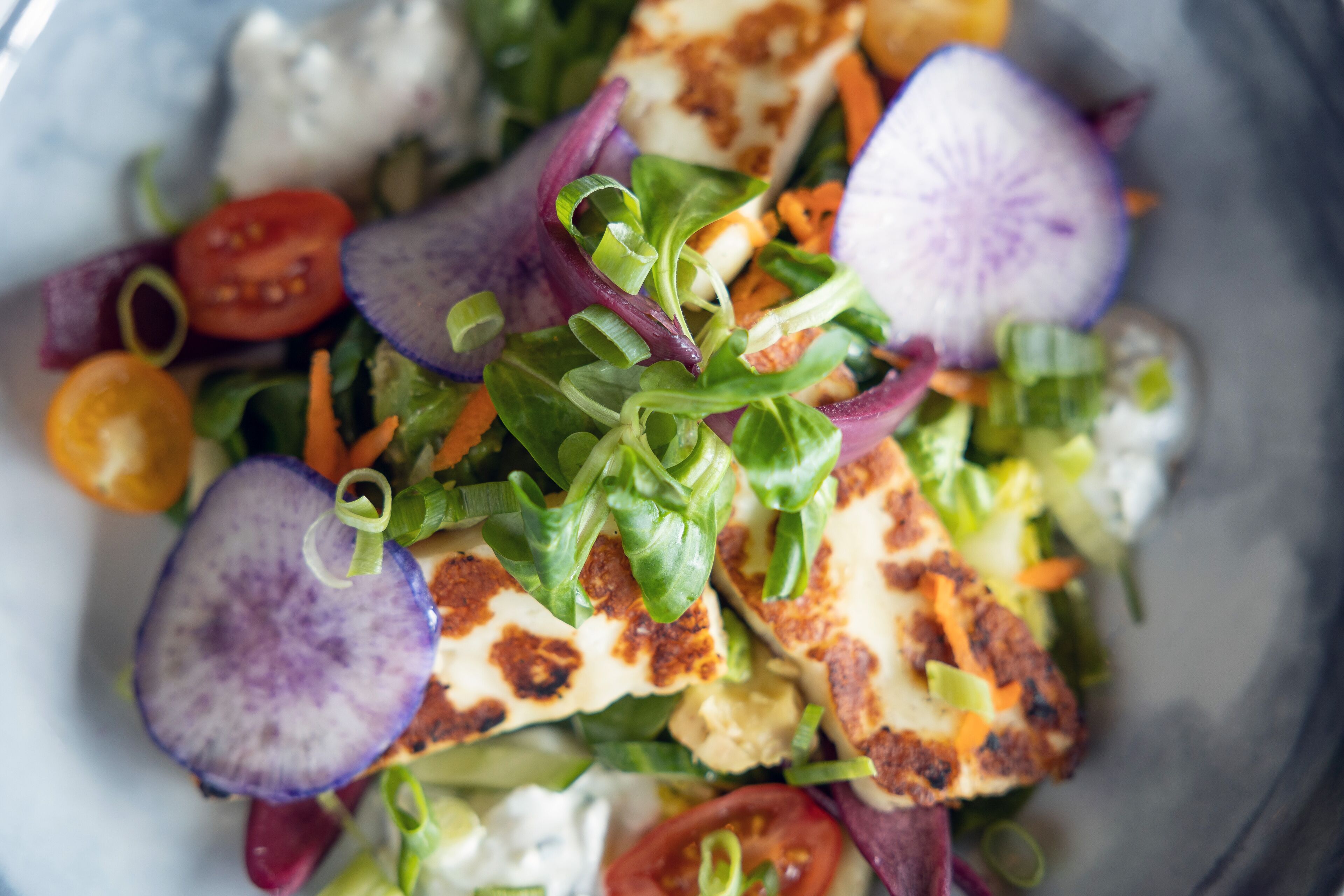 Primer plano de una ensalada con diversas verduras y un trozo de queso a la plancha.