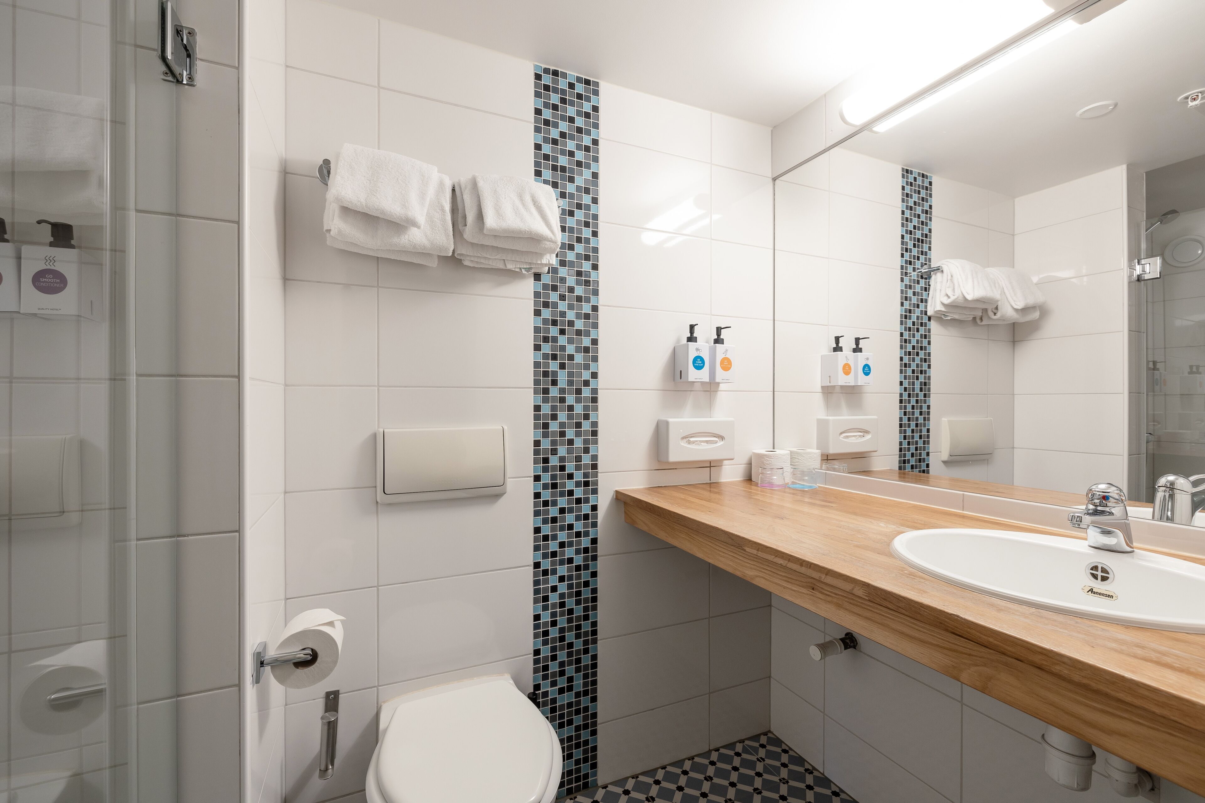 Un baño moderno con inodoro blanco, lavabo con encimera de madera y espejo. Hay dos secadores de manos y un dispensador de toallas de papel en la pared.