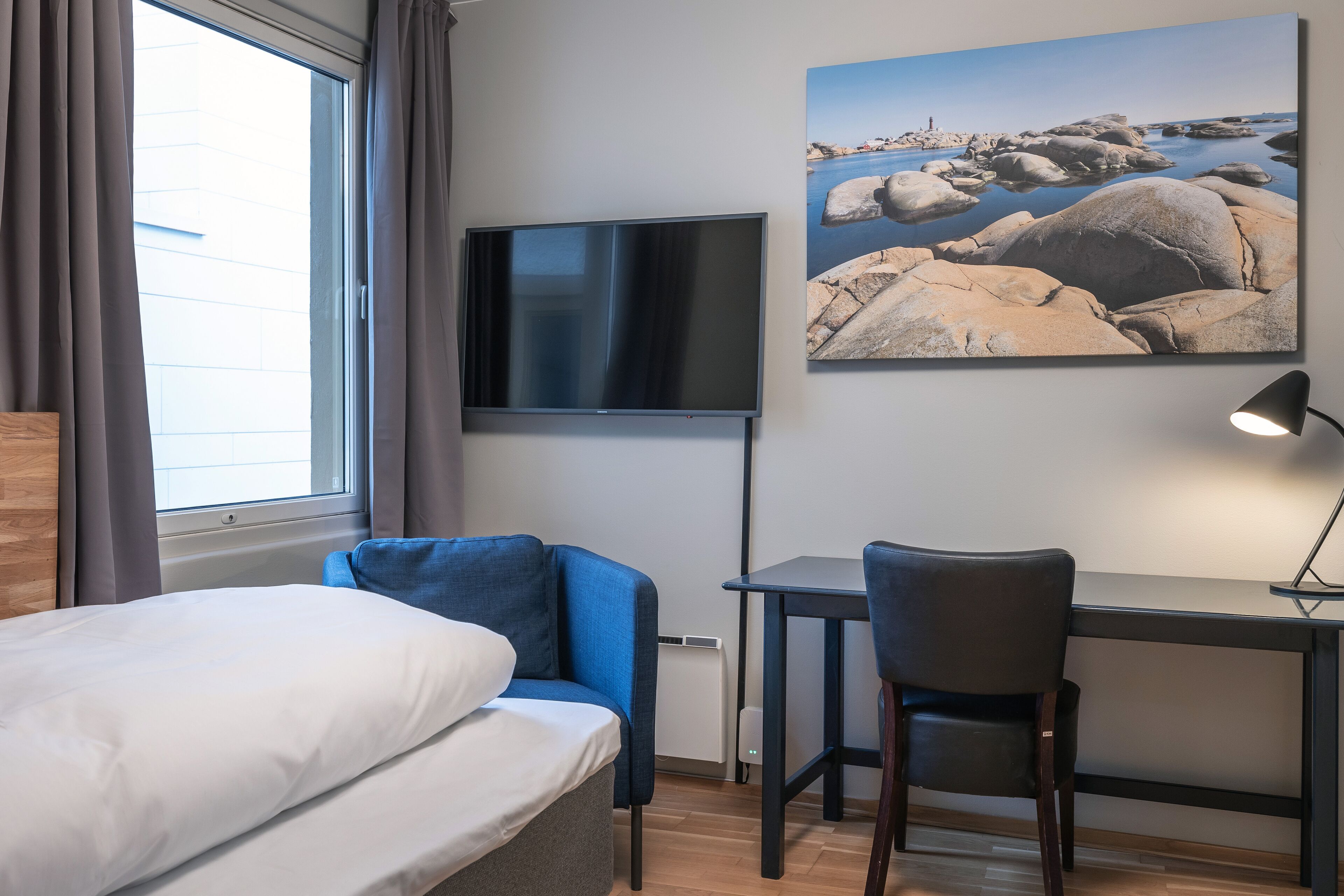 A hotel room with a bed, a desk, a chair, a TV, and a painting of a rocky landscape.