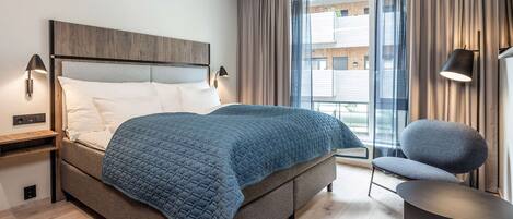 Una habitación de hotel moderna con una cama grande, una silla, una mesita y una ventana con cortinas.