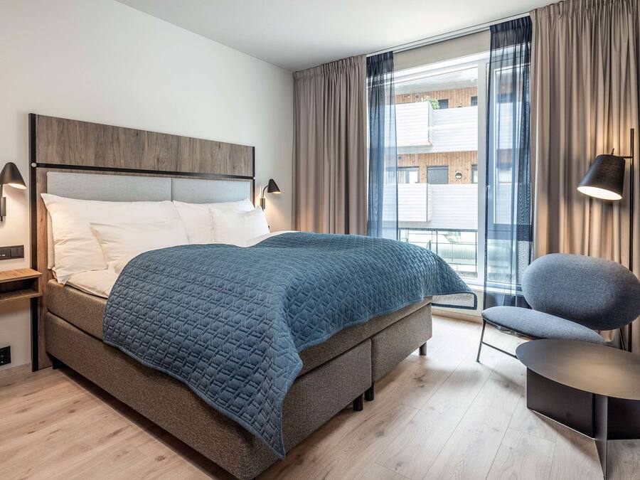 Une chambre d’hôtel moderne avec un grand lit, une chaise, une petite table et une fenêtre avec des rideaux.