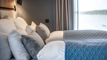 A neatly made bed with multiple pillows and a blue quilt, a wall-mounted lamp, and a view of a body of water through a window with curtains.