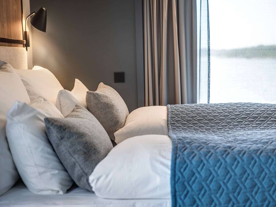 A neatly made bed with multiple pillows and a blue quilt, a wall-mounted lamp, and a view of a body of water through a window with curtains.