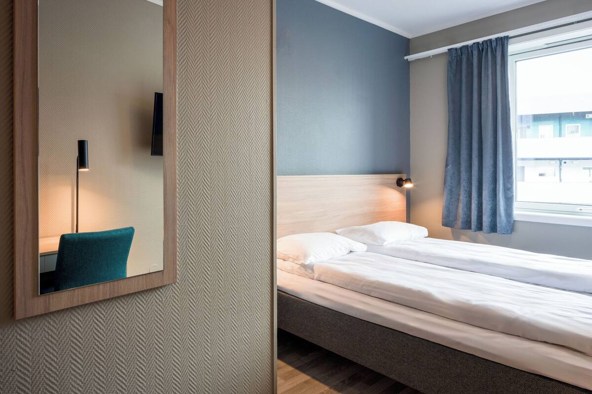 A hotel room with a bed, a bedside table, a mirror, and a window with curtains.