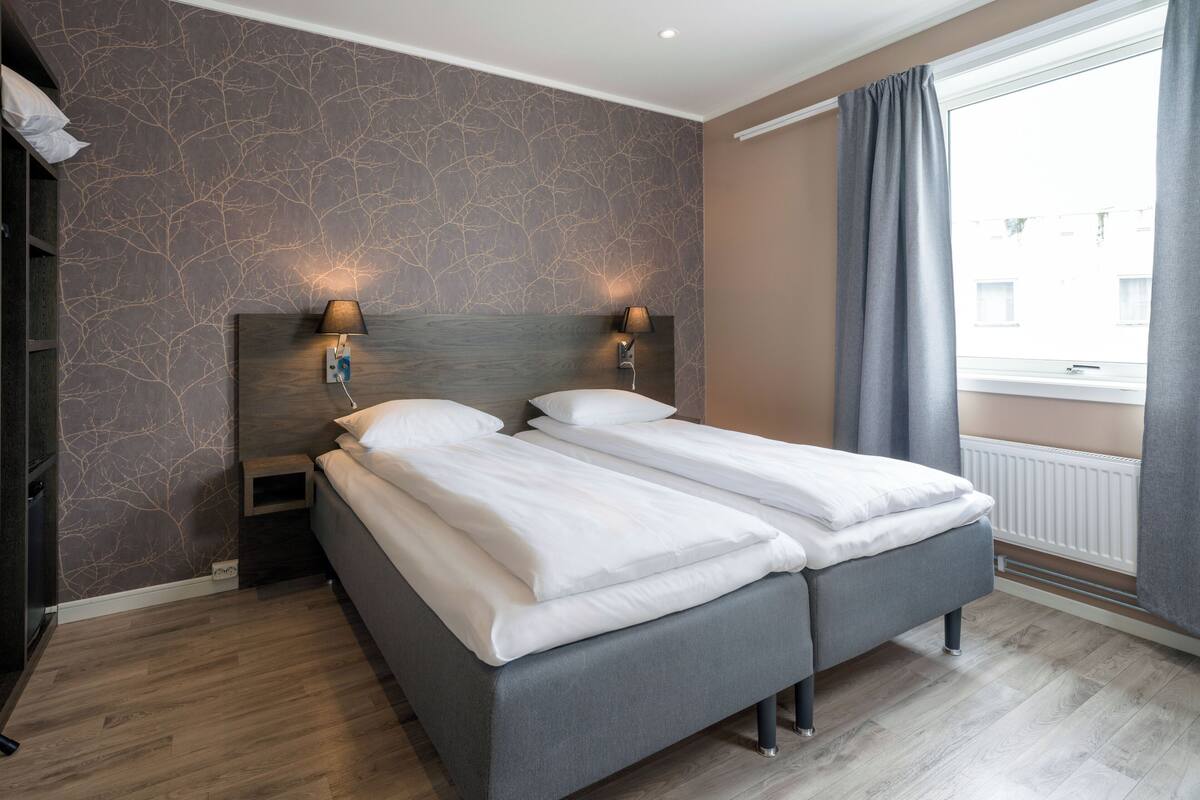 A double bed with two bedside tables, each with a lamp, in a room with a patterned wall and a window with curtains.