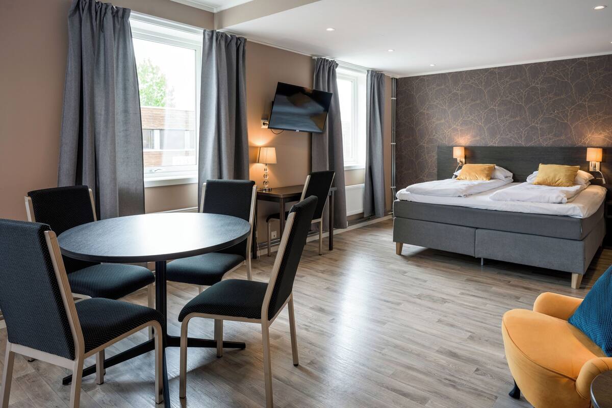 A modern hotel room with a bed, a desk, a round table with chairs, and a TV.