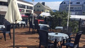 An outdoor patio with wooden flooring, tables, and chairs, surrounded by buildings with visible signage.