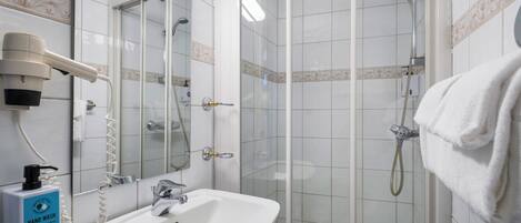 Un baño con un lavamanos blanco, un secador de manos, una ducha con cerramiento de vidrio y una toalla colgada en la pared.