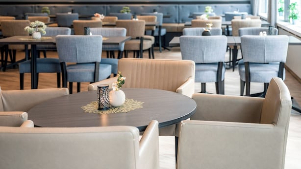 A modern restaurant with round tables and cushioned chairs, framed pictures on the wall, and natural light from large windows.