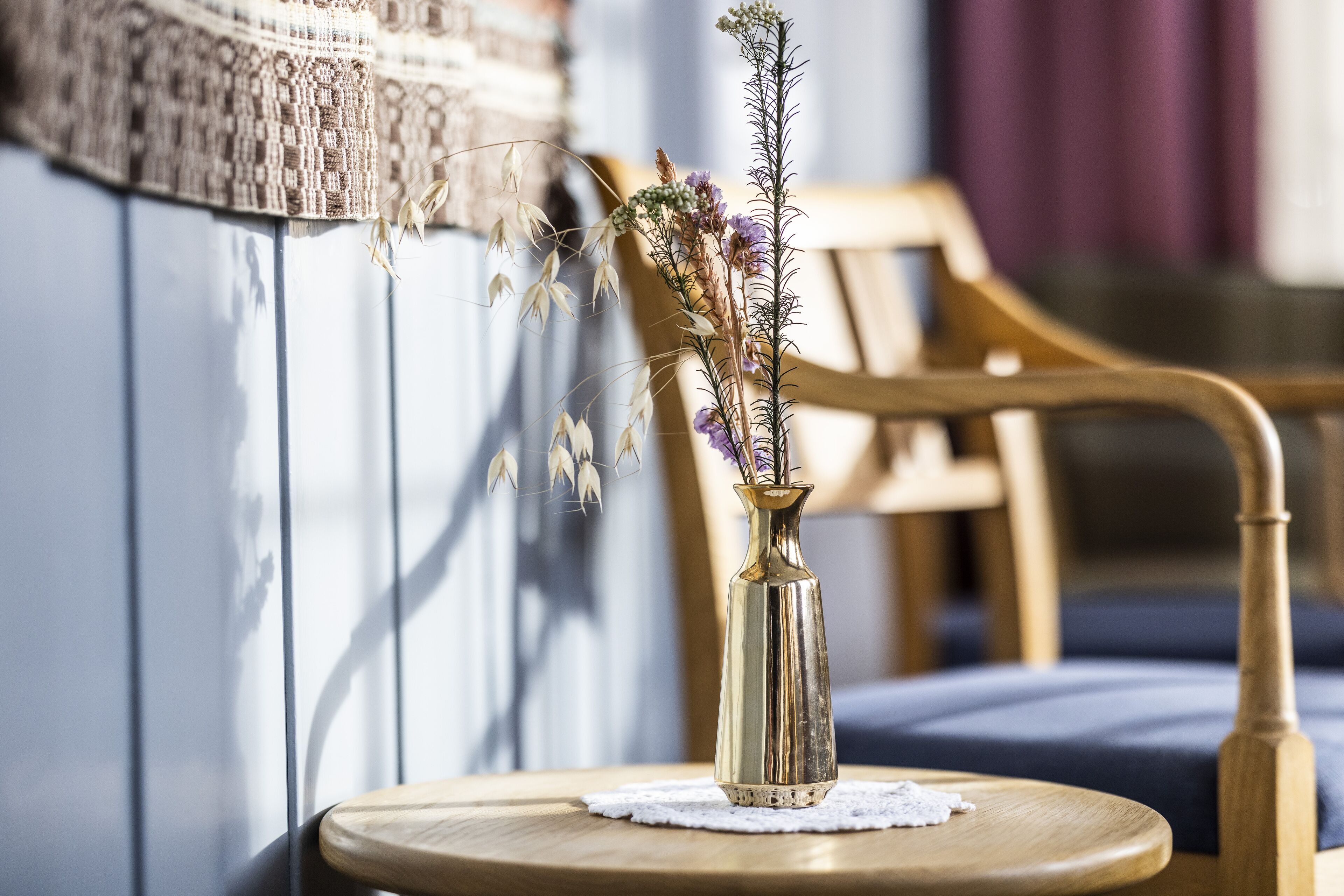 Una silla de madera con cojín, una mesita redonda pequeña con un mantel de encaje y un jarrón dorado con flores secas.