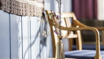 A wooden chair with a cushion, a small round table with a lace doily, and a gold vase with dried flowers.