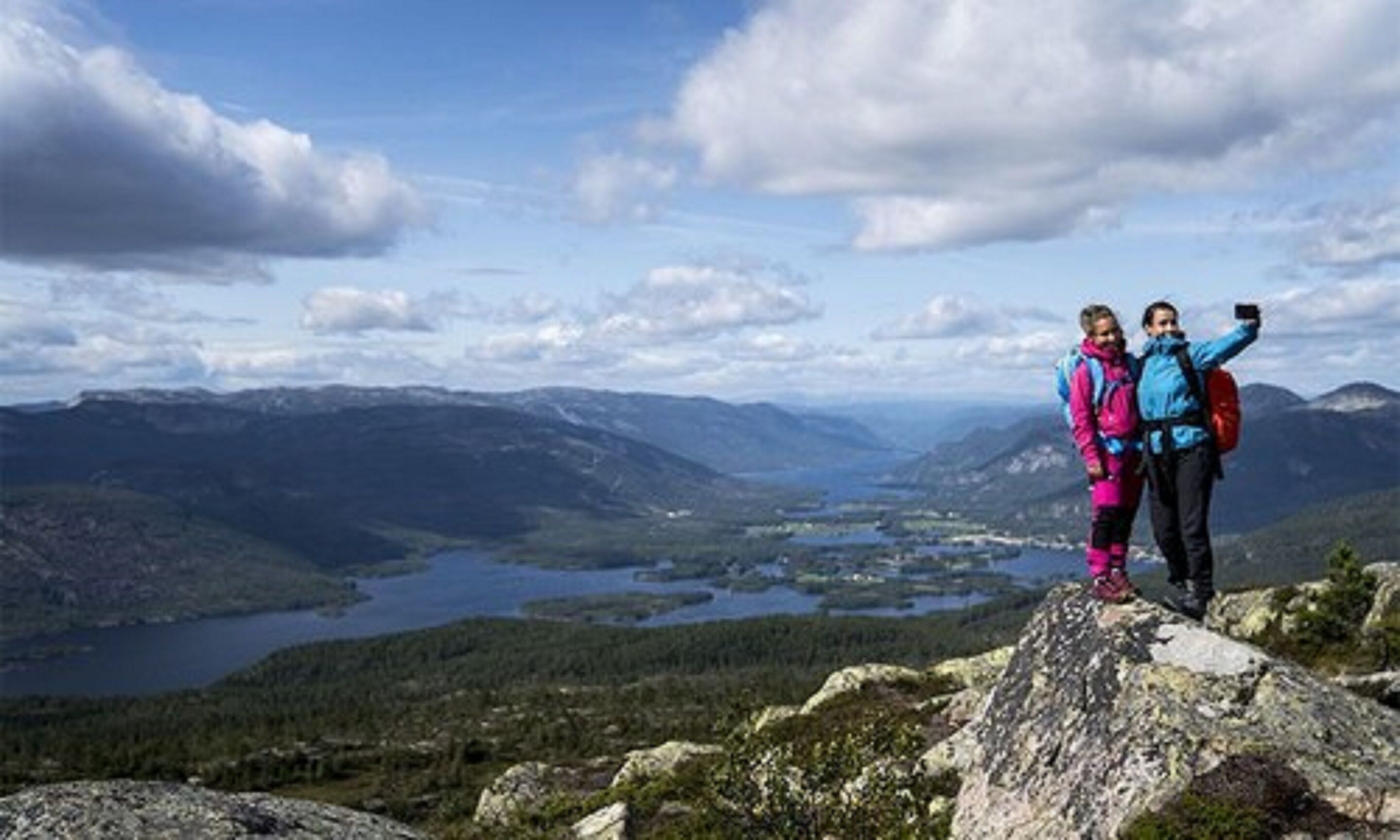 Två personer står på en klippavsats med ett bergigt landskap och en sjö i bakgrunden.