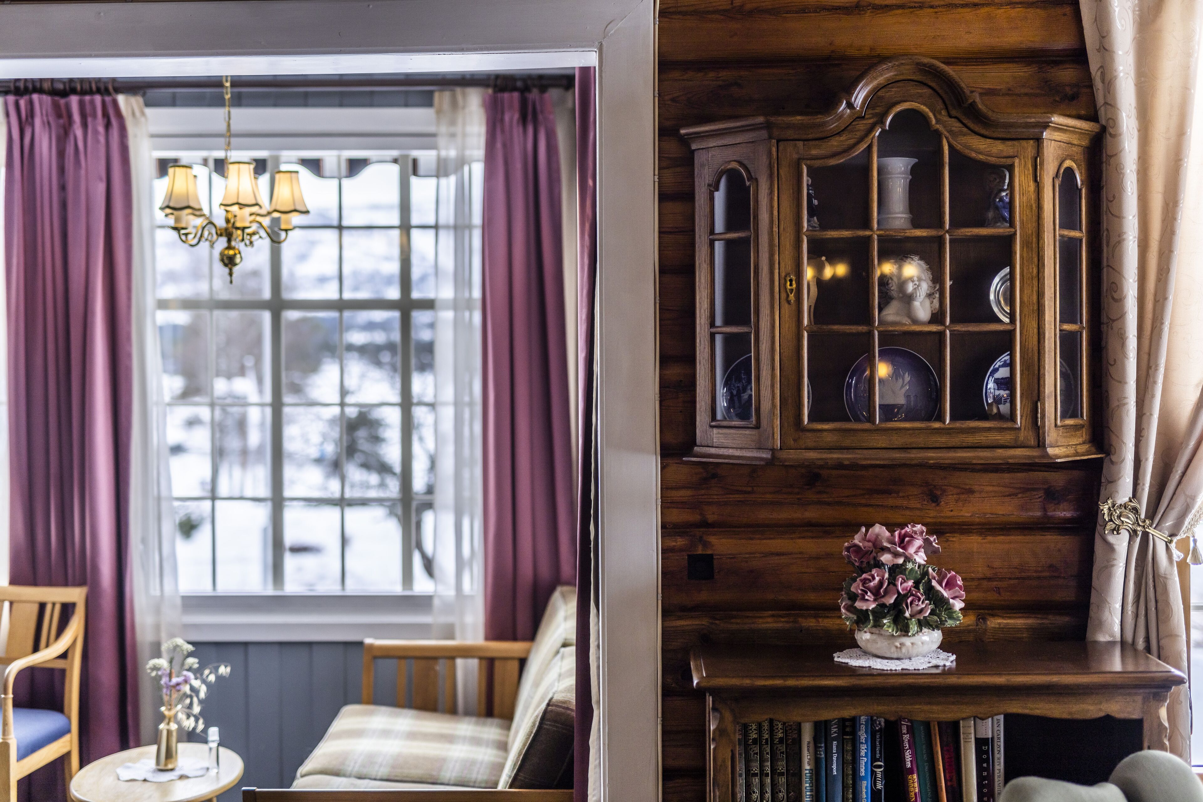 Una habitación acogedora con ventana, un aparador de madera con puertas de cristal y una pequeña mesa con un florero.