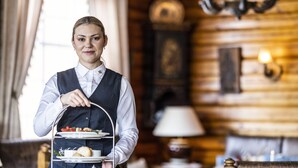 A person in a uniform holding a three-tiered tray with food items, in a room with wooden walls and a lamp. - Straand Hotel (Kviteseid)