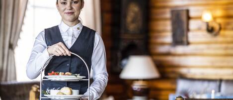 A person in a uniform holding a three-tiered tray with food items, in a room with wooden walls and a lamp.