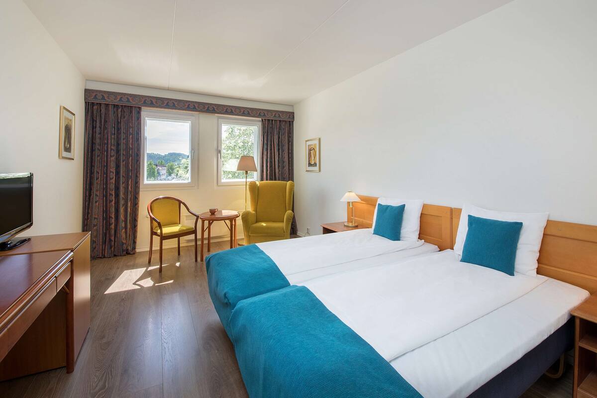 Habitación de hotel con una cama grande, un escritorio con televisor, una silla, una mesita y una ventana con cortinas.