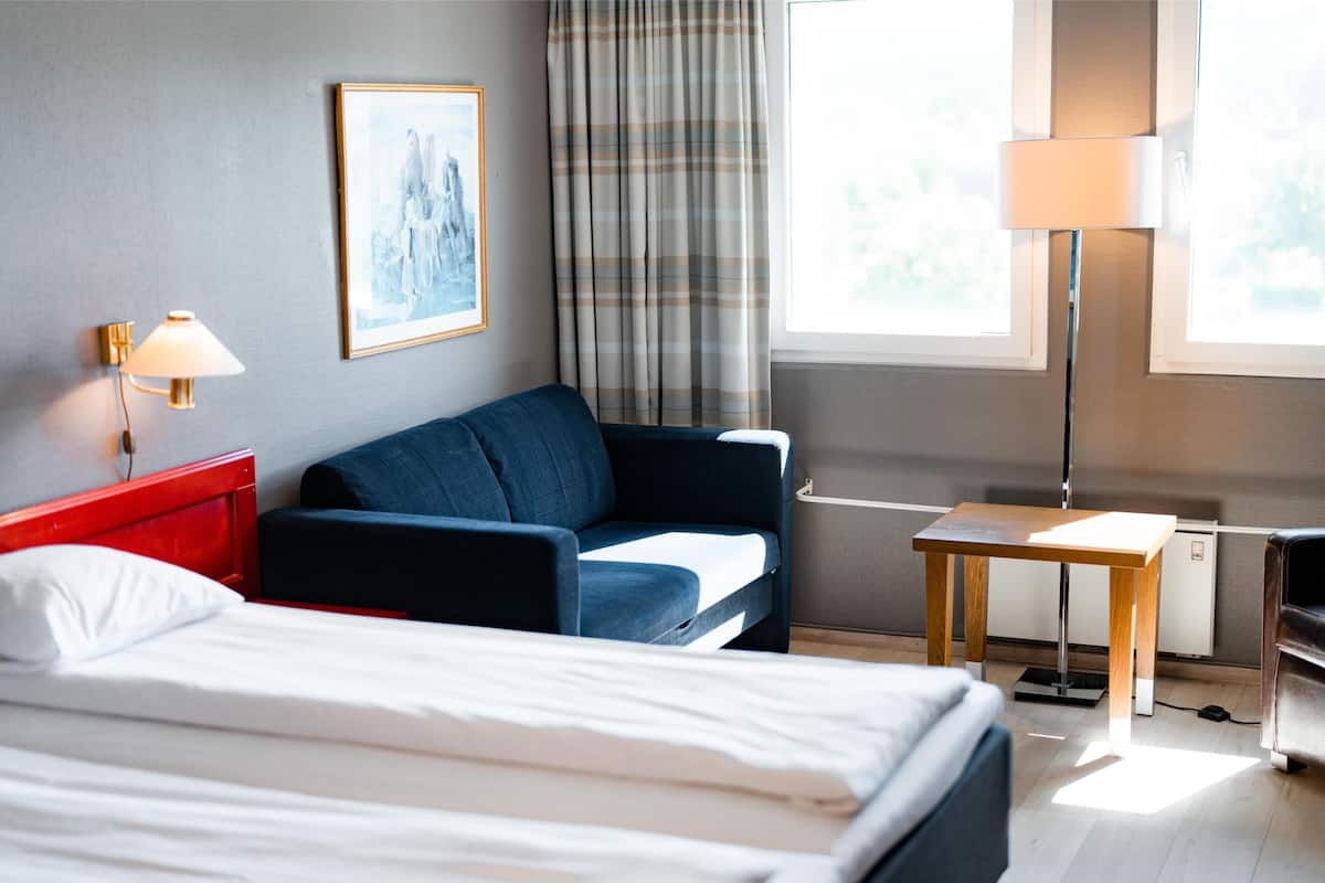 Habitación de hotel con cama, sofá, una mesita, lámpara y ventana con cortinas.