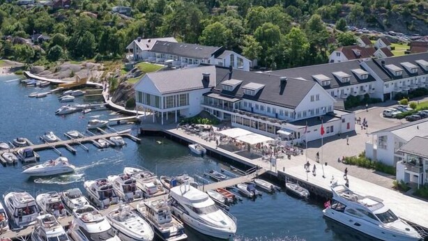 A marina with multiple boats docked, a large building with a sloped roof, and a hilly landscape in the background. - Havna Hotel & Sjøbad (Færder)