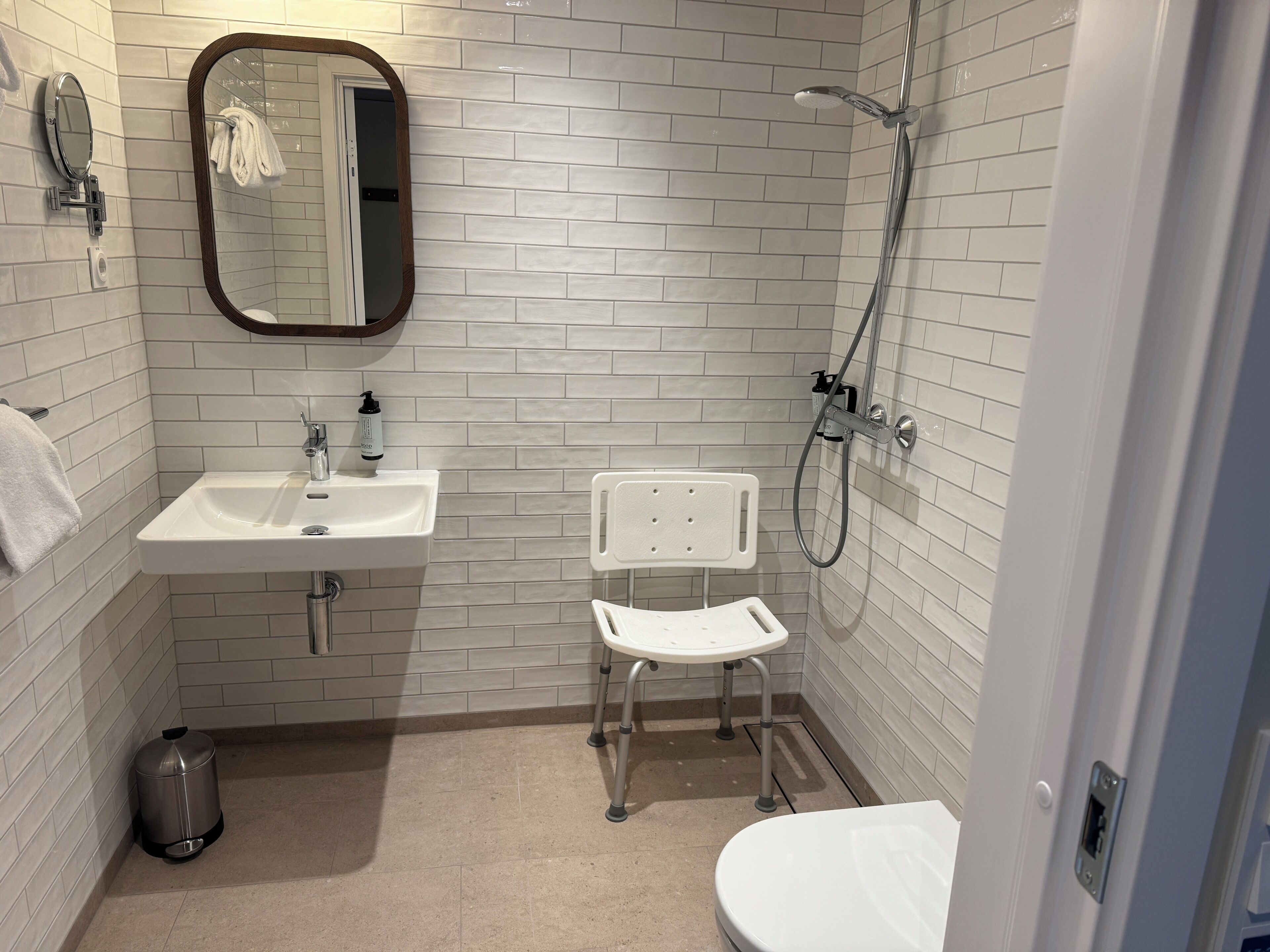 Un bany amb parets de rajola blanca, un lavabo, un mirall, una dutxa i un vàter.