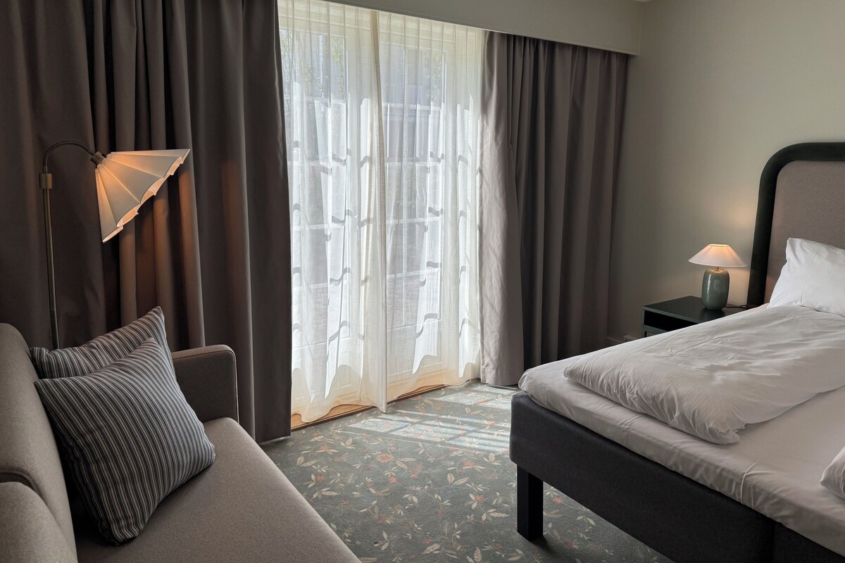 A hotel room with a bed, a sofa, a lamp, a nightstand, and a view through the curtains.