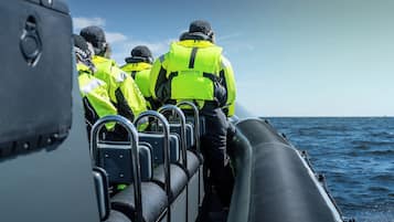 A group of people in high-visibility jackets on a boat, with metal railings and a view of the sea.