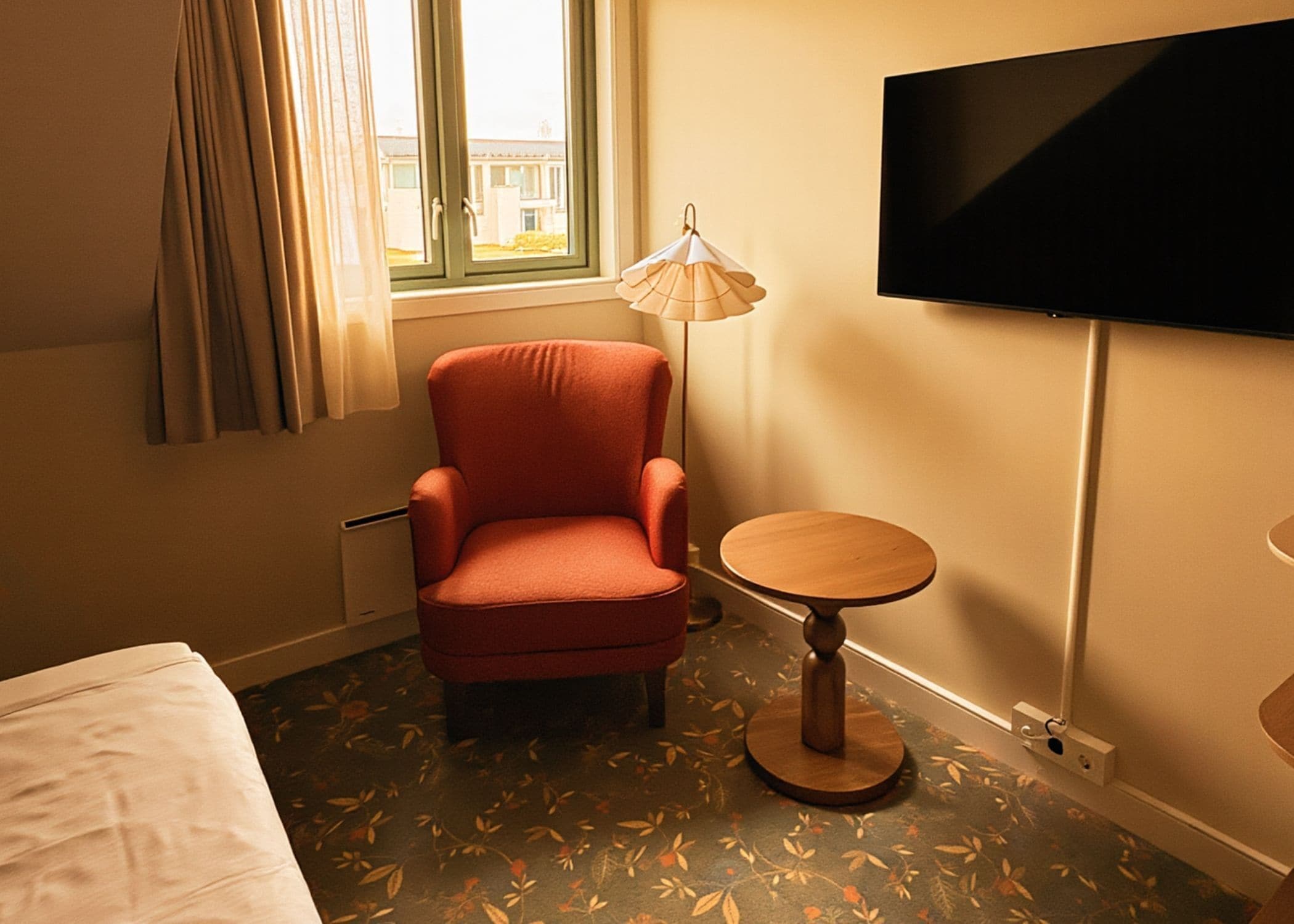 Et hotelværelse med en enkelt seng, en rød lænestol, et lille rundt bord, en gulvlampe og et fjernsyn monteret på væggen.