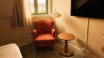 Et hotelværelse med en enkelt seng, en rød lænestol, et lille rundt bord, en gulvlampe og et fjernsyn monteret på væggen.