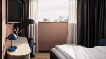 A hotel room with a bed, a desk with a lamp, a chair, a TV mounted on the wall, and a window with curtains.