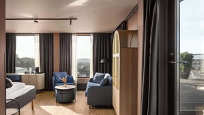A modern hotel room with a bed, a sofa, a chair, a small table, and a wardrobe. Large windows offer a view of the city. - Scandic Sarpsborg (Sarpsborg)