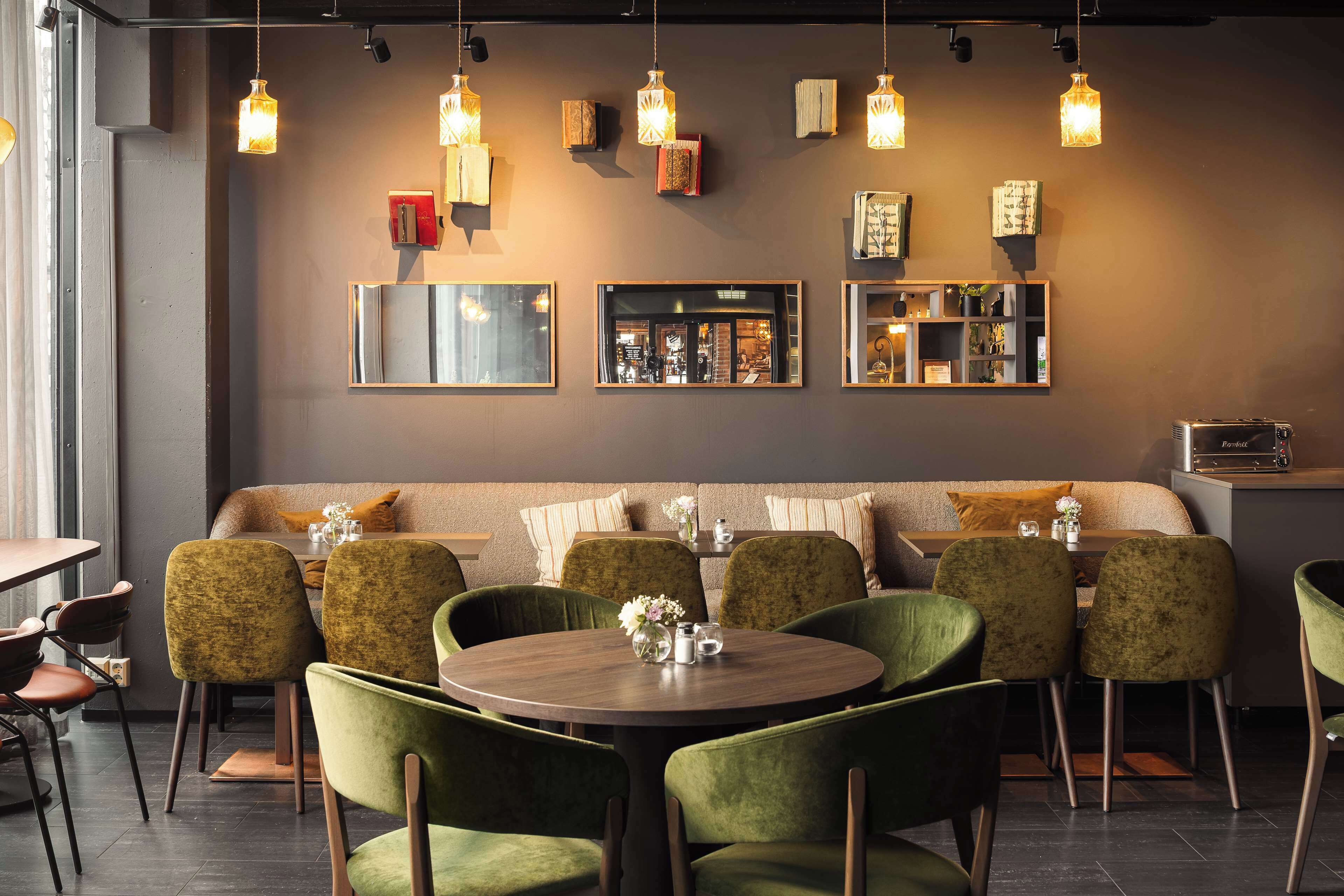 A restaurant interior with a round table, green upholstered chairs, a sofa, hanging pendant lights, and framed artwork on the walls.