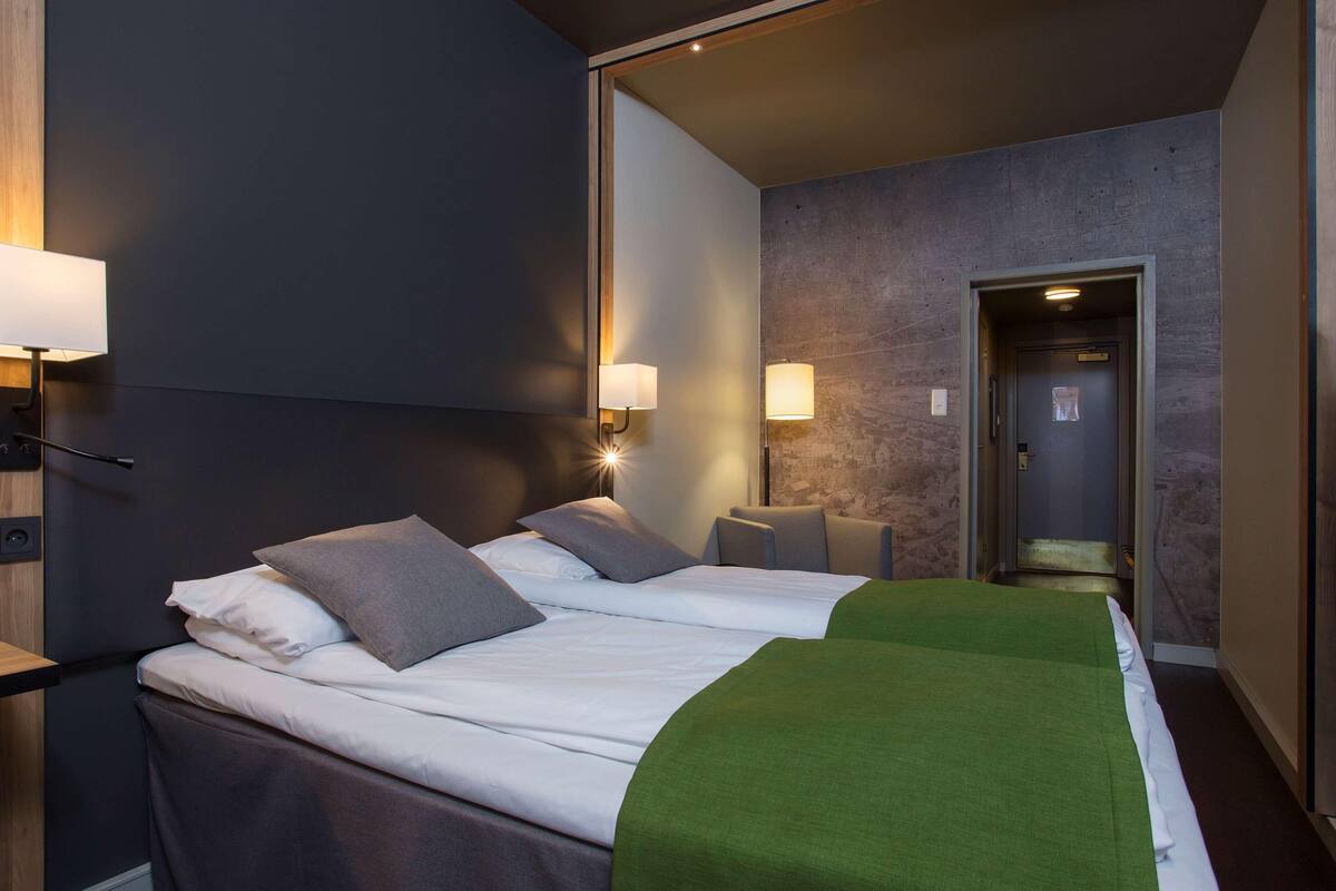 A modern hotel room with a large bed, two bedside lamps, and a green throw blanket.