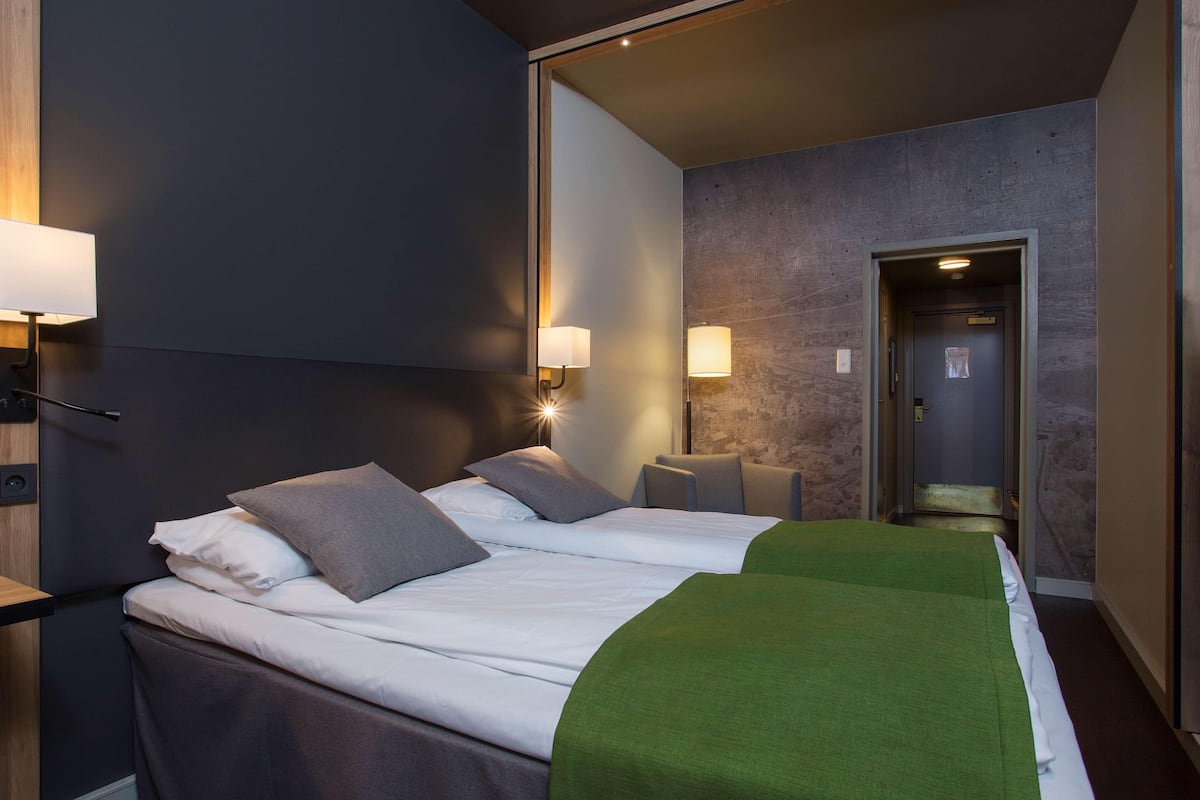 A modern hotel room with a large bed, two bedside lamps, a green throw blanket, and a door leading to another room.