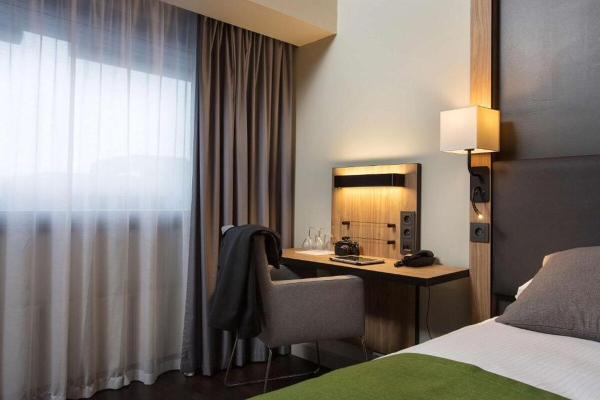 A hotel room with a bed, a desk with a chair, a lamp, a mirror, and a window with curtains.