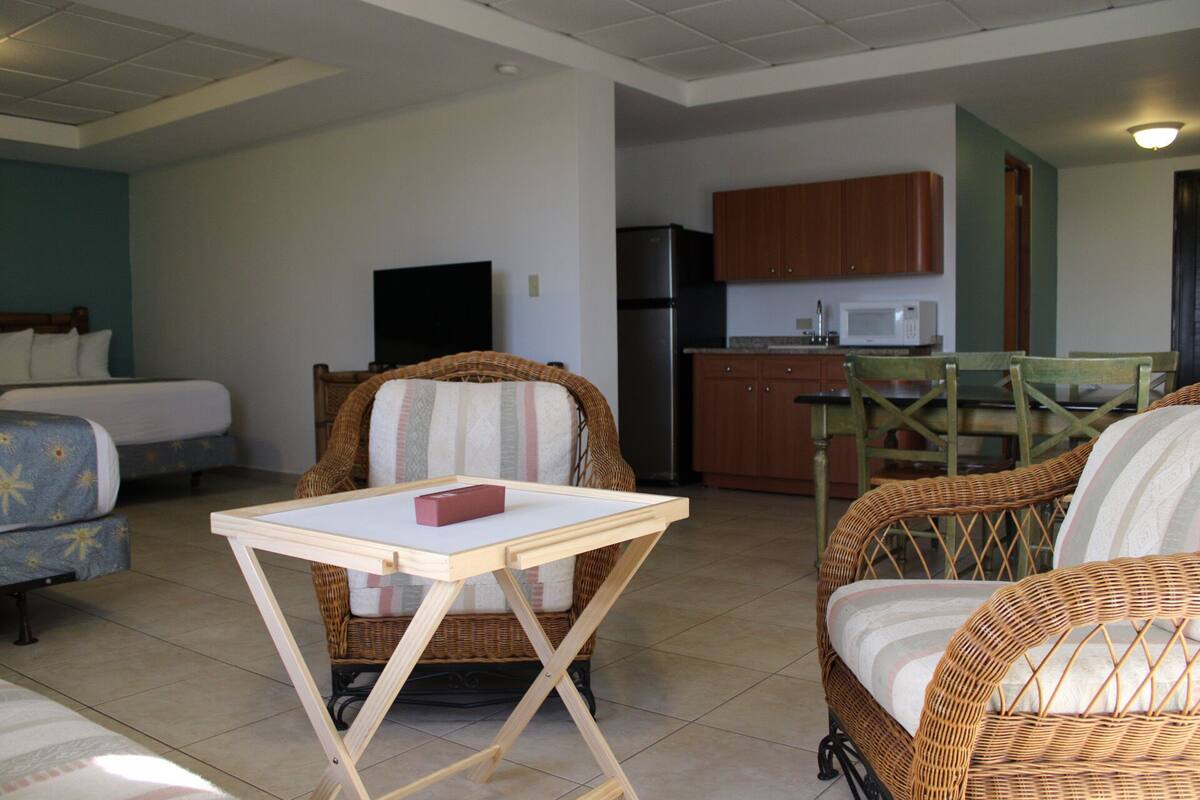 A room with a bed, a wicker chair, a small table, and a kitchenette in the background.