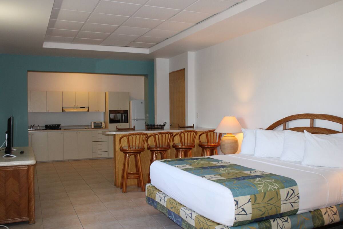 A hotel room with a bed, a desk, a chair, a small kitchenette, and a bathroom.
