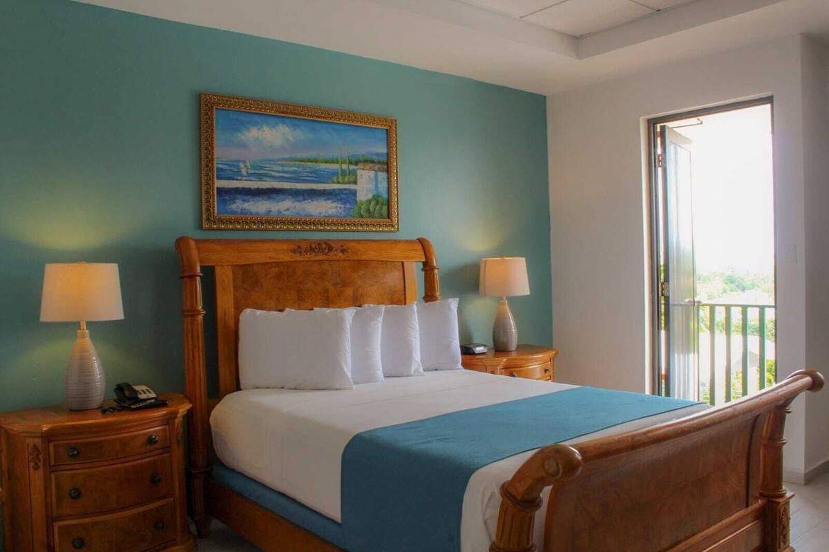 A bedroom with a wooden bed, two bedside tables, a painting above the bed, and a view of the outdoors through a window.