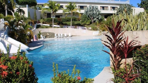 Une piscine à l’eau d’un bleu limpide, entourée de plantes tropicales, avec un bâtiment visible au loin.