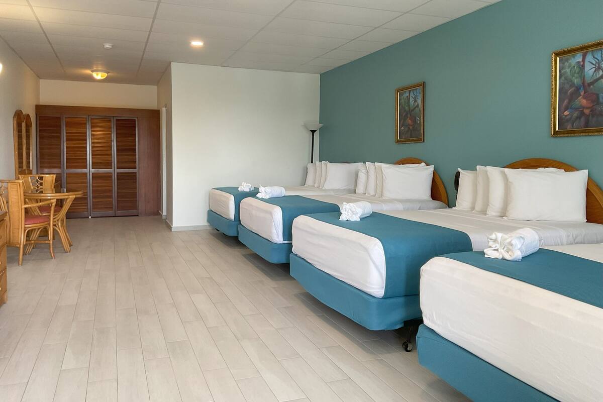 A hotel room with four single beds, a wooden dining table with chairs, a wardrobe, and a painting on the wall.