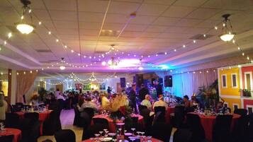 A banquet hall with round tables covered in red tablecloths, string lights, and a band performing on a raised platform.